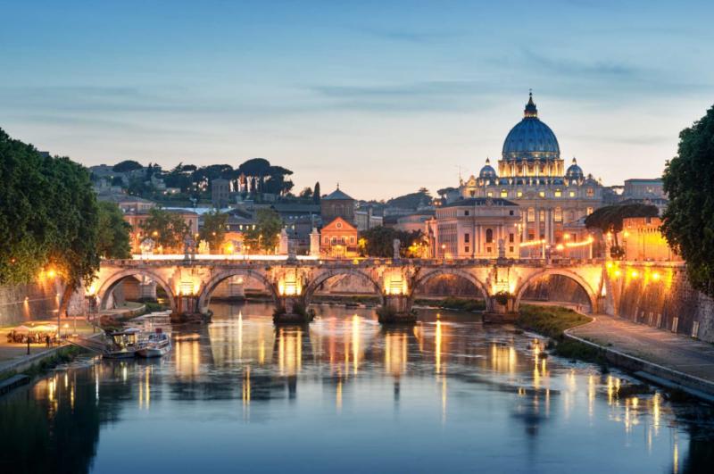 a bridge and buildings with their lights on with a river at dusk